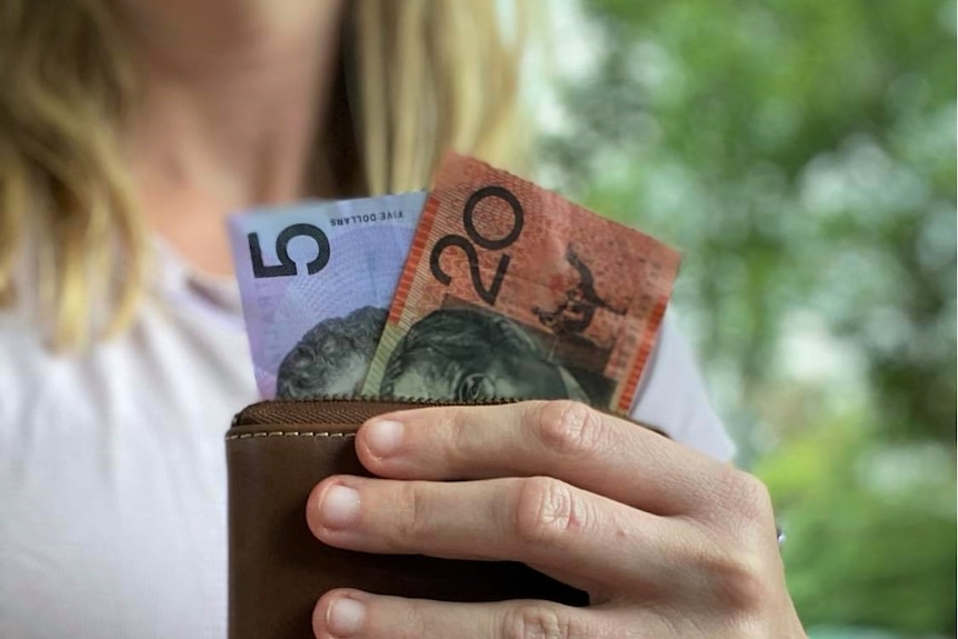 A woman holds a leather wallet with a $5 and $20 note in it.