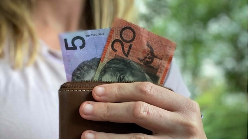A woman holds a leather wallet with a $5 and $20 note in it.