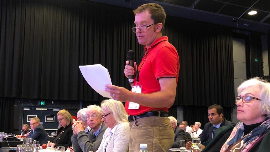 Man standing at conference table in audience of auditorium speaking and holding a piece of paper