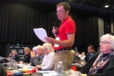 Man standing at conference table in audience of auditorium speaking and holding a piece of paper