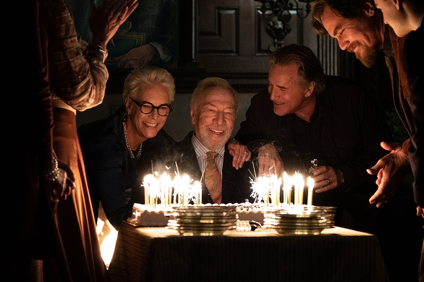 Jamie Lee Curtis, Christopher Plummer and Don Johnson smile and are illuminated by birthday cake sparklers in dark room.