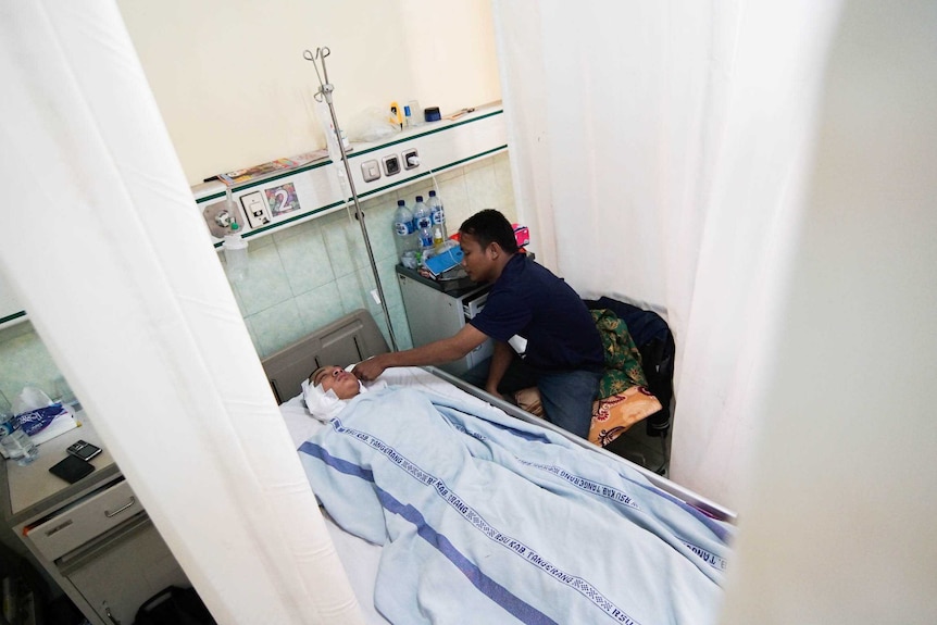 A man leans over his young brother lying in a hospital bed
