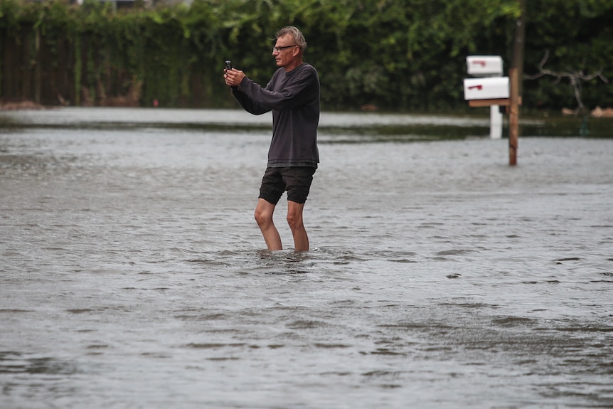 A man up to his midcalfs in flood water taking a photo. 