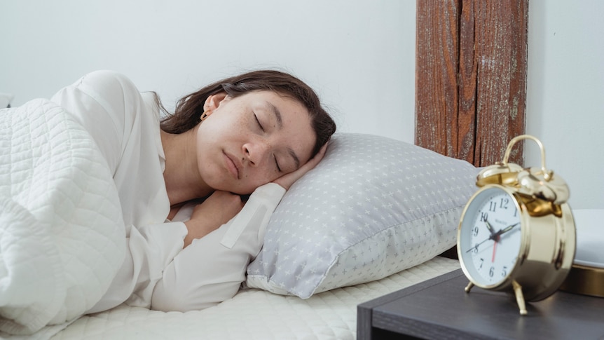 A woman with dark brown hair sleeping in a bed, made in white linen, next to a gold alarm clock on a bedside table