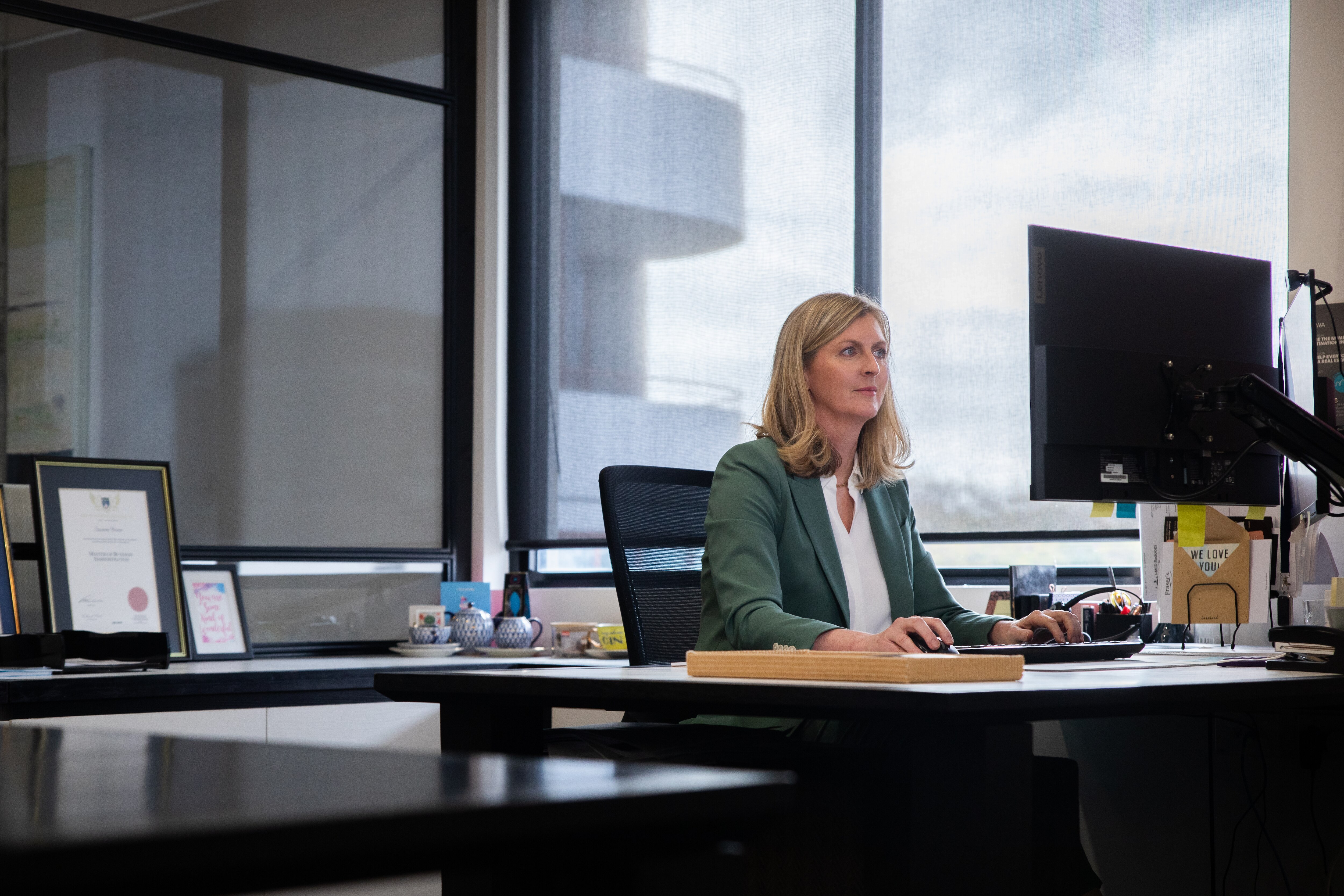 A woman with blonde hair in a green jacket sits at her computer in front of a large window.