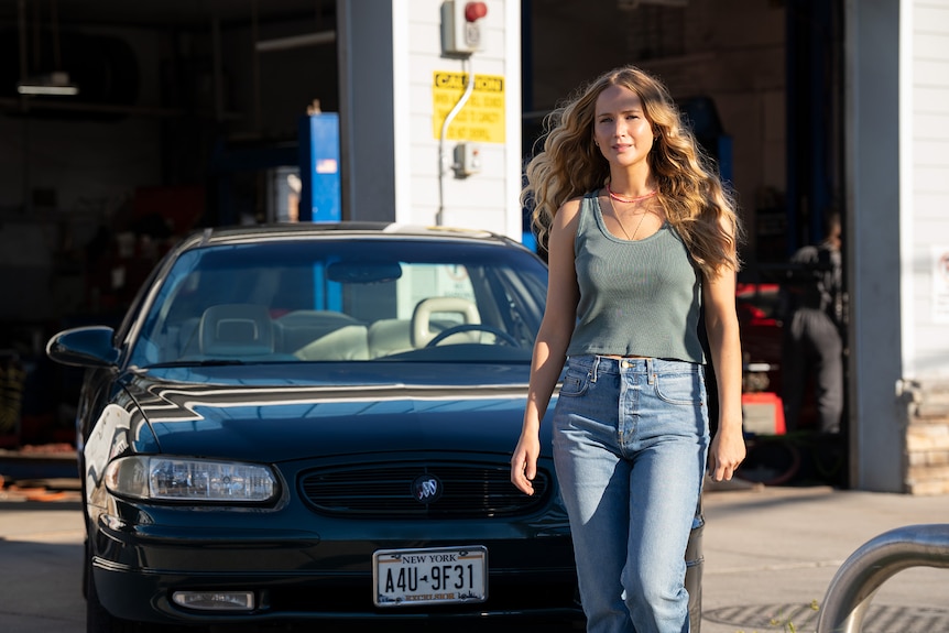 A blonde white woman wearing a khaki singlet and blue jeans walks in front of a car at a mechanic shop.