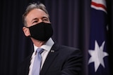 Greg Hunt, wearing a black face mask, stands in front of a blue curtain and an Australian flag
