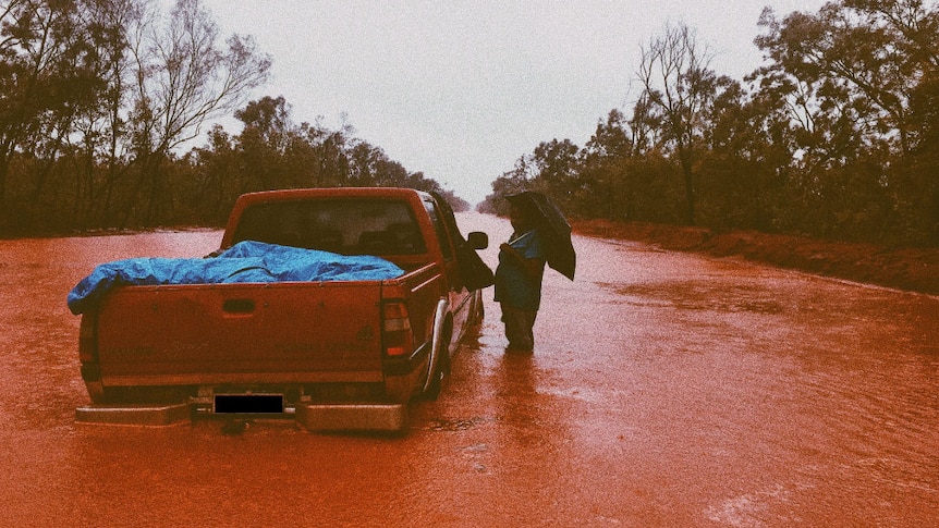 A person holding an umbrella standing knee-deep in red floodwater next to a car on a flooded road.