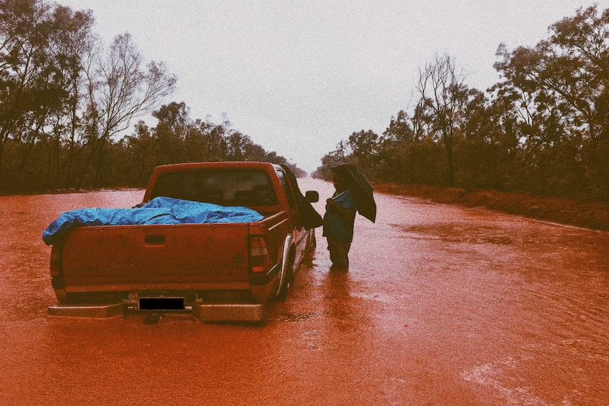 A person holding an umbrella standing knee-deep in red floodwater next to a car on a flooded road.