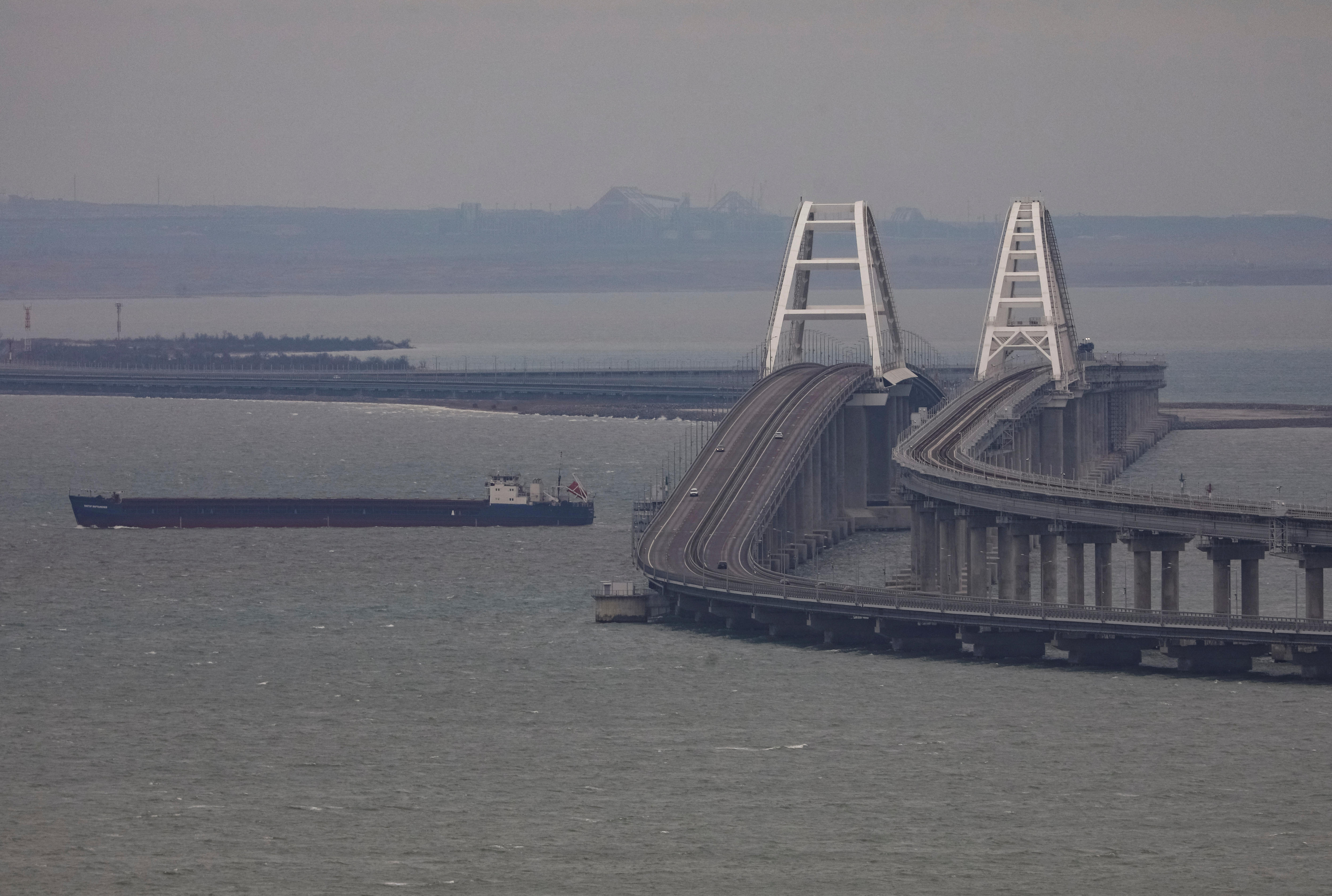 A barge floats next to a long double-span bridge which curves back and forth over a grey body of water.