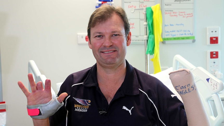 A man in a West Coast Eagles t-shirt sits on a bed holding up his hands, one with two fingers missing and another bandaged