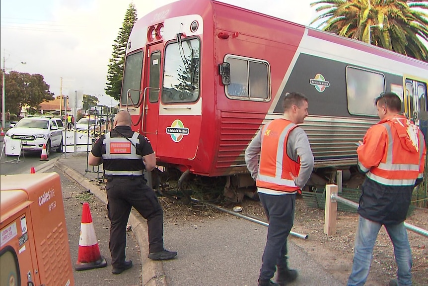 Three people in orange vests stand in front of a red train that has overshot the track