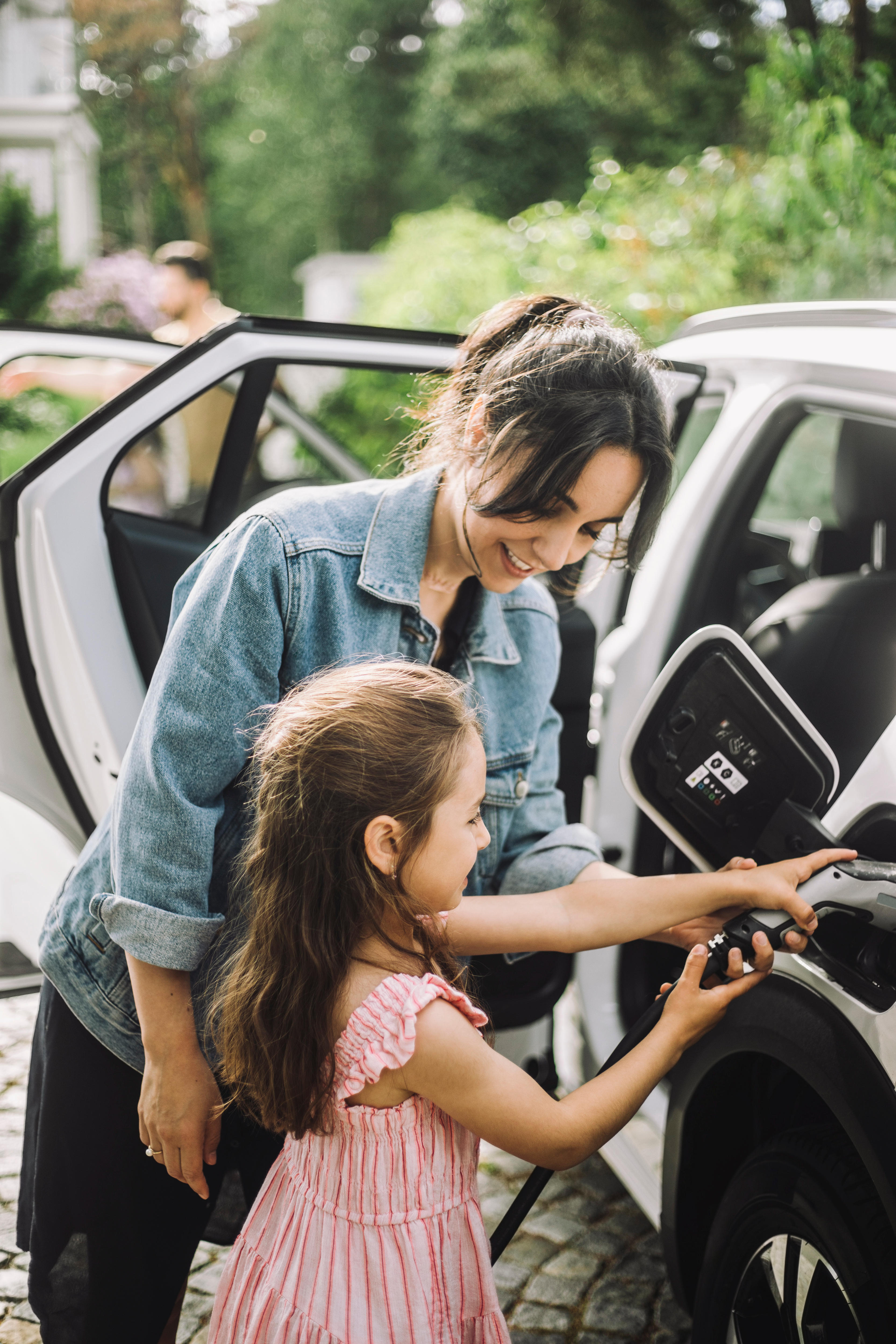 A dark-haired woman in a denim jacket helps a child charge a white electric car.