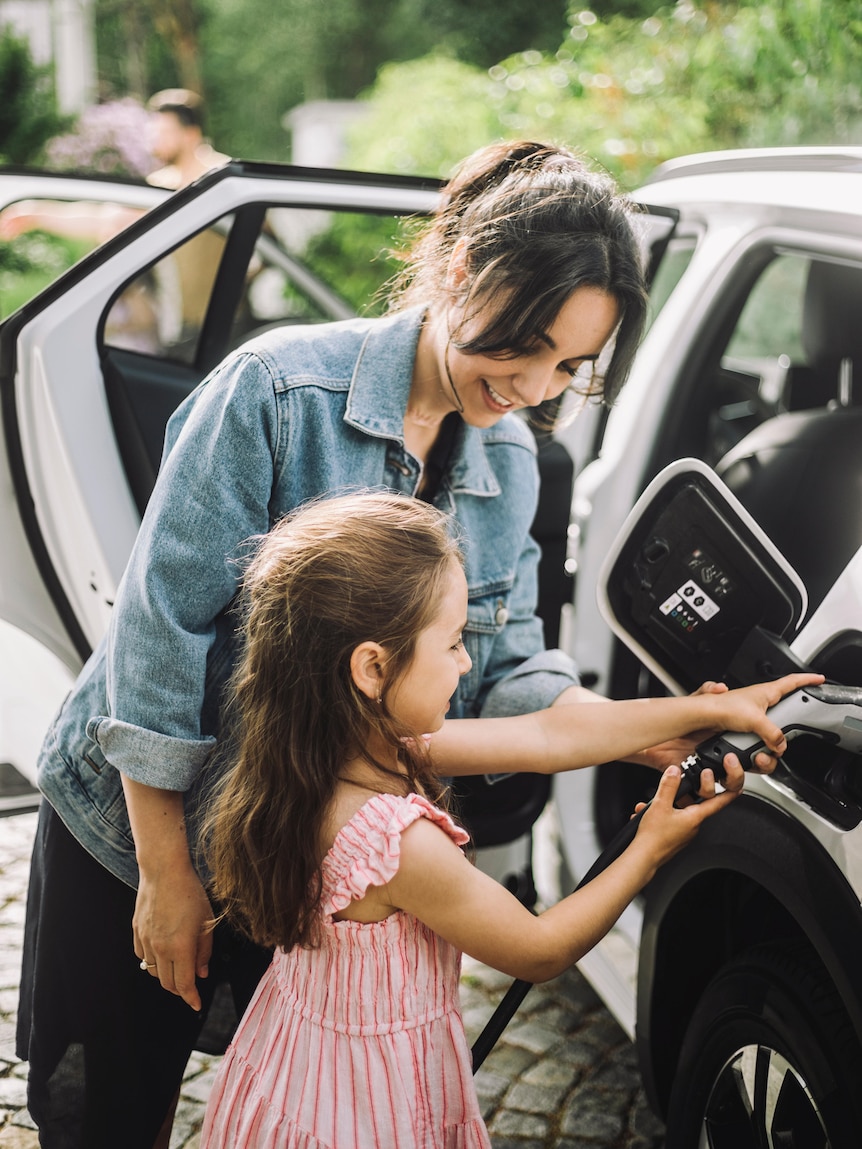 A dark-haired woman in a denim jacket helps a child charge a white electric car.