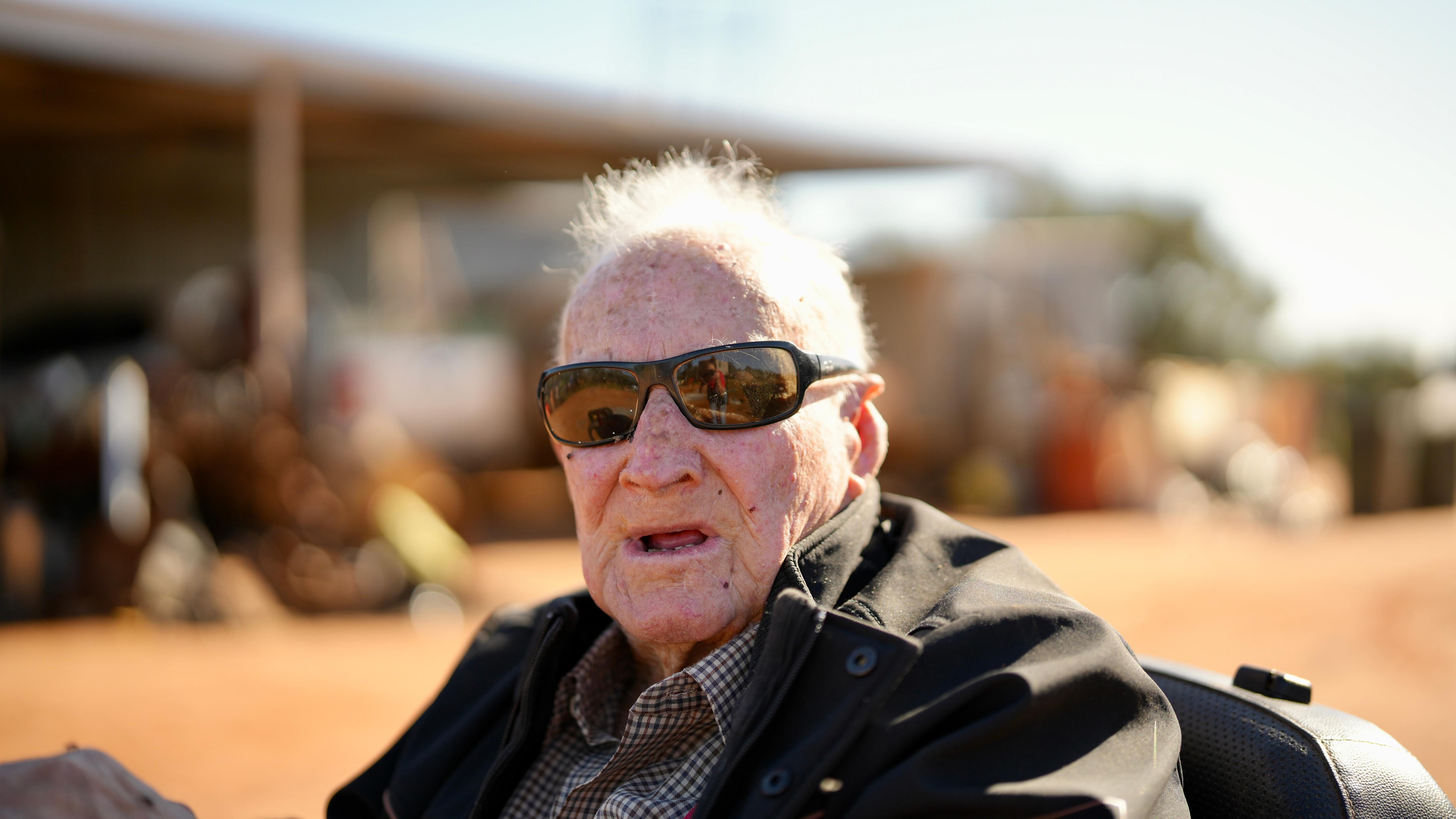 an old man with black sunglasses and a red face close to the camera looking 