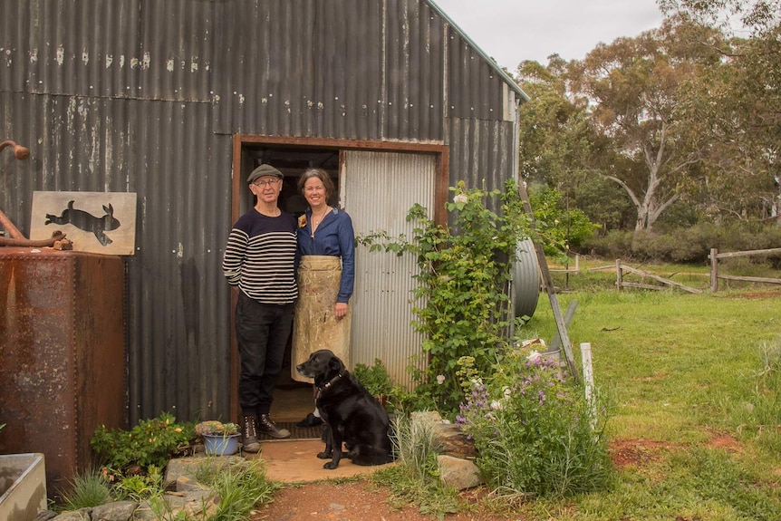 A man, woman and dog outside an old tin shed in a garden with bushland in the background