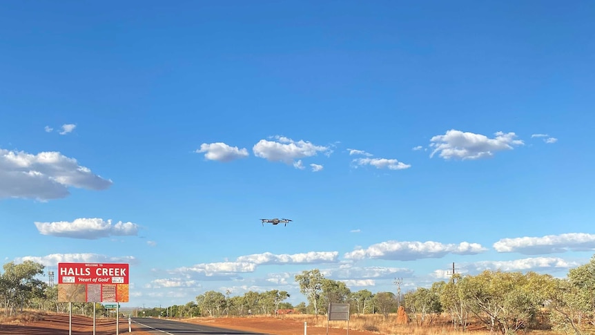 A small flying machine hovers above the highway near a signpost for Halls Creek