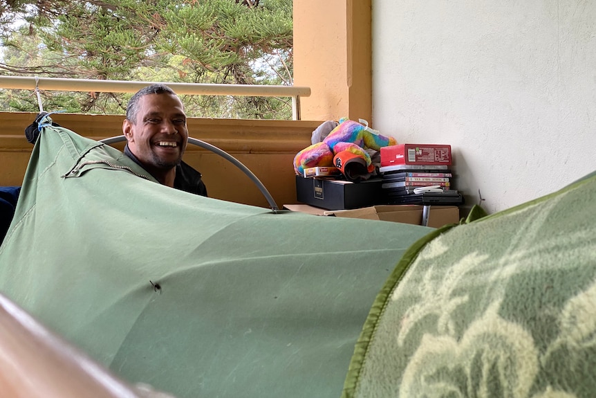 A man smiles at the camera from his green swag with a pile of his belongings behind him