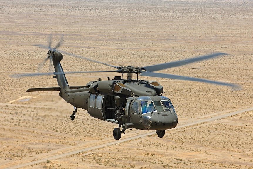 A green military helicopter flies above yellow dry, desolate land.  