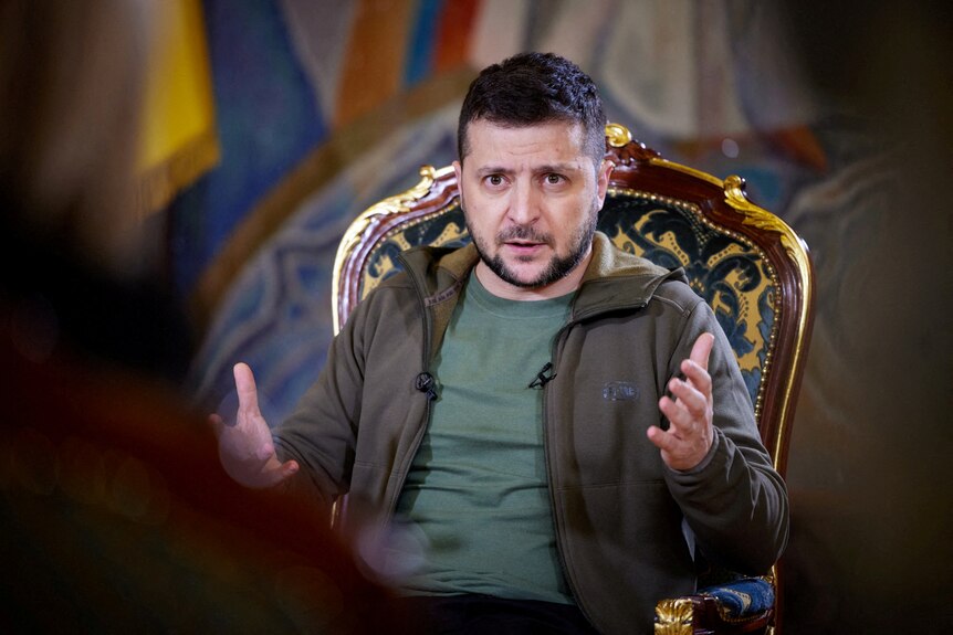 Volodomyr Zelenskyy, wearing a khaki green T-shirt under a darker hoodie, gestures while sitting in an ornate chair 