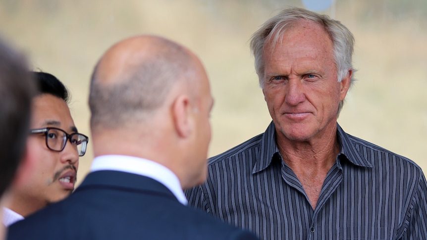 A man with white hair and a navy striped shirt talks to a bald man whose back is to the camera.