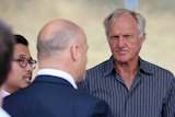A man with white hair and a navy striped shirt talks to a bald man whose back is to the camera.