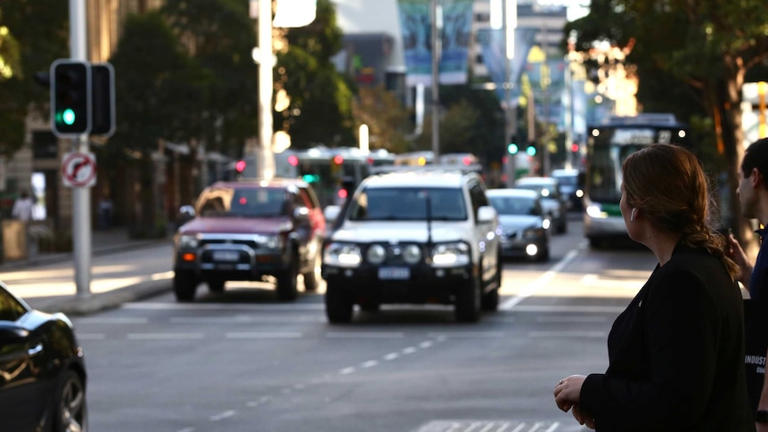 A woman waits at a pedestrian crossing in the CBD as cars drive past.