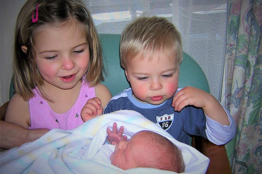 Two kids, a girl about 4 and a boy about 1 ogling over a swaddled newborn baby.