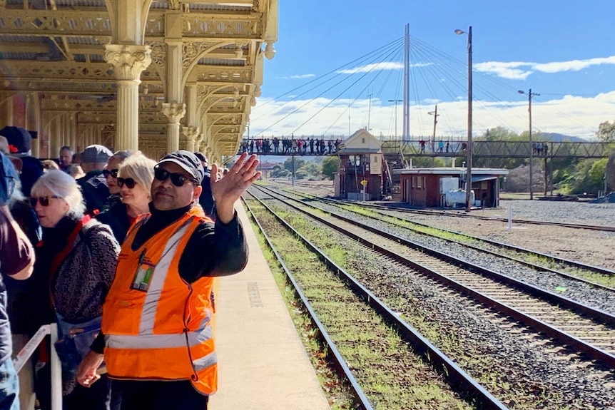 Crowds gather on the platform and the overpass at a railway station on a sunny day.