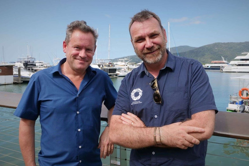 Two men standing in front of a railing at a marina with boats in the background.