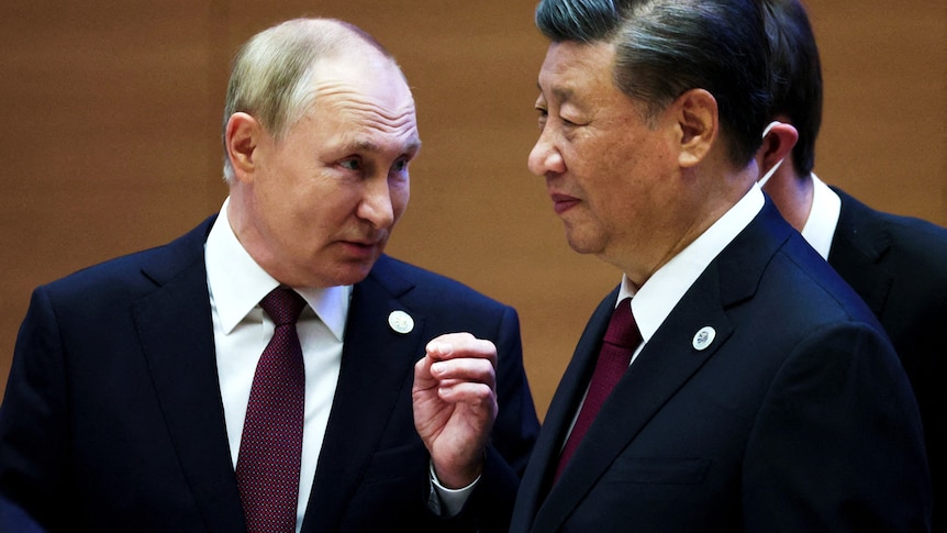 Vladimir Putin dressed in a suit and tie looks over at Xi Jinping dressed in a suit.