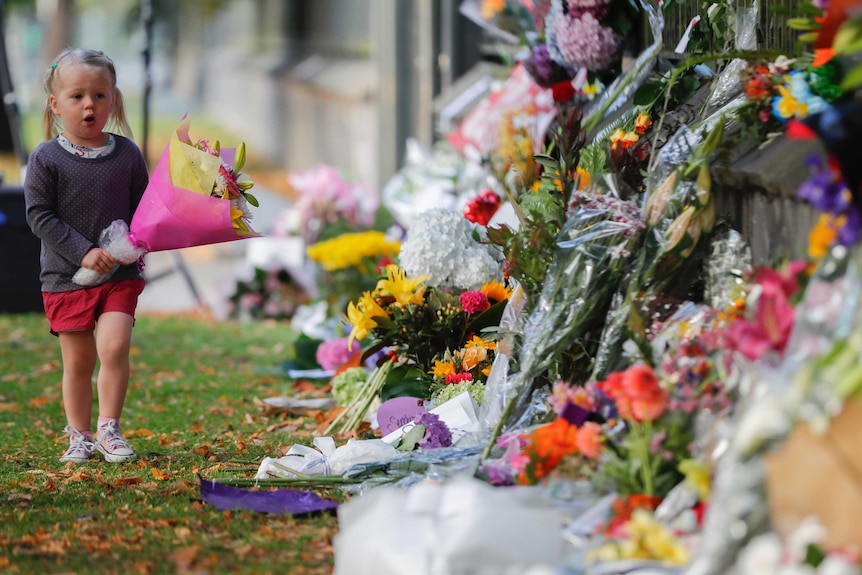 A small blonde girl carries a bunch of flowers to a wall with many bunches of flowers.