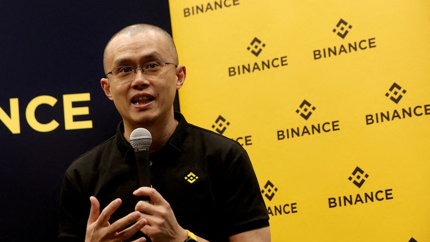 A bald man of Chinese heritage wearing glasses and a black polo shirt holds a microphone in front of a yellow backdrop.