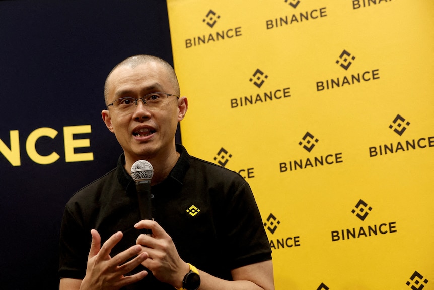 A bald man of Chinese heritage wearing glasses and a black polo shirt holds a microphone in front of a yellow backdrop.