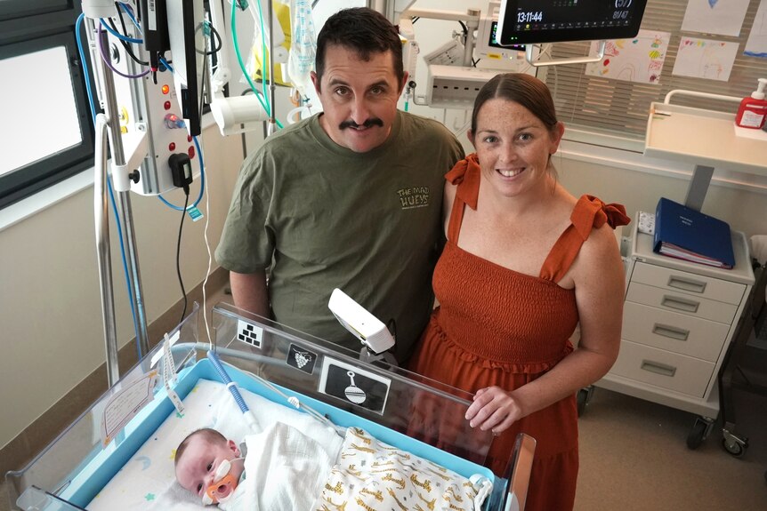 A man and a woman stand next to a baby in a cot in a hospital room