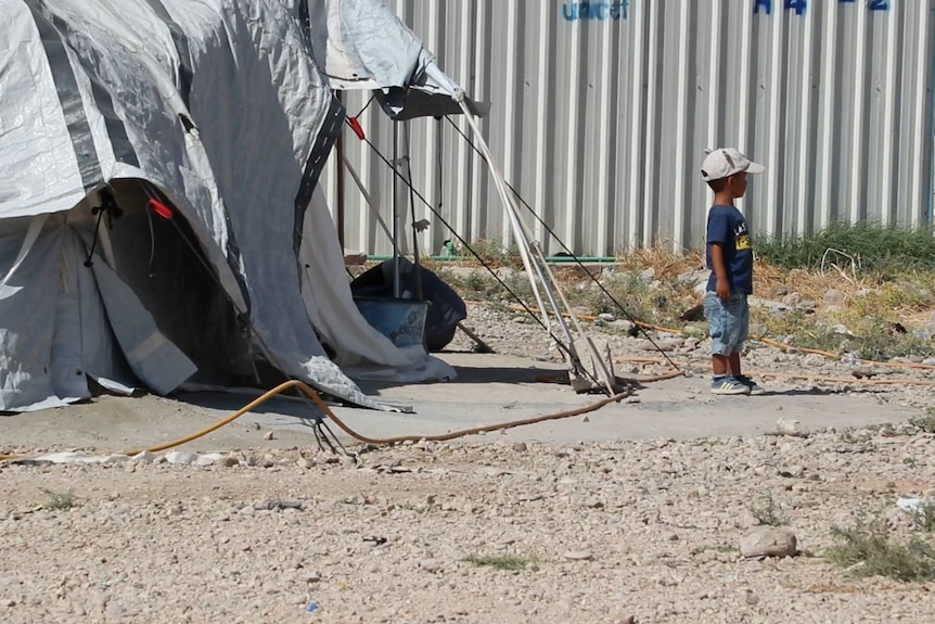 A small child stands outside a tent.