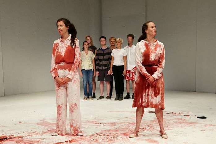 Two women covered in blood stand in front of a group of people in casual clothes on stage