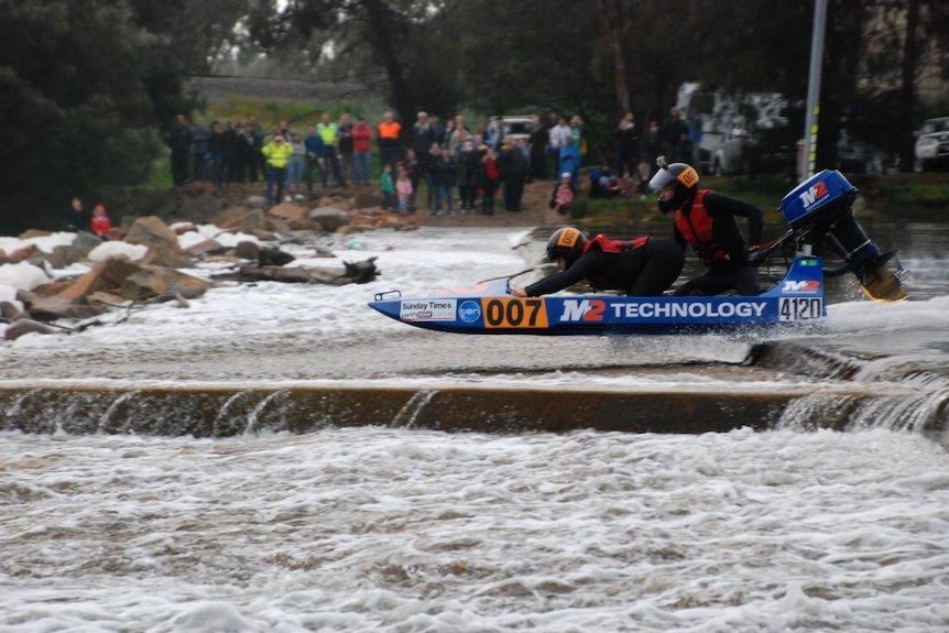 A blue powerboat with two people onboard is driven forward on the Avon River in front of spectators.