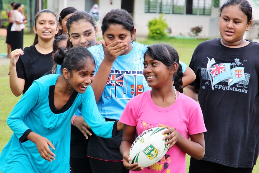 A group of girls is laughing and talking to one girl holding a rugby league ball.