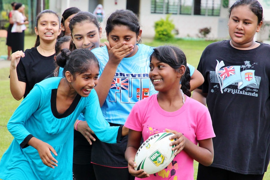 A group of girls is laughing and talking to one girl holding a rugby league ball.