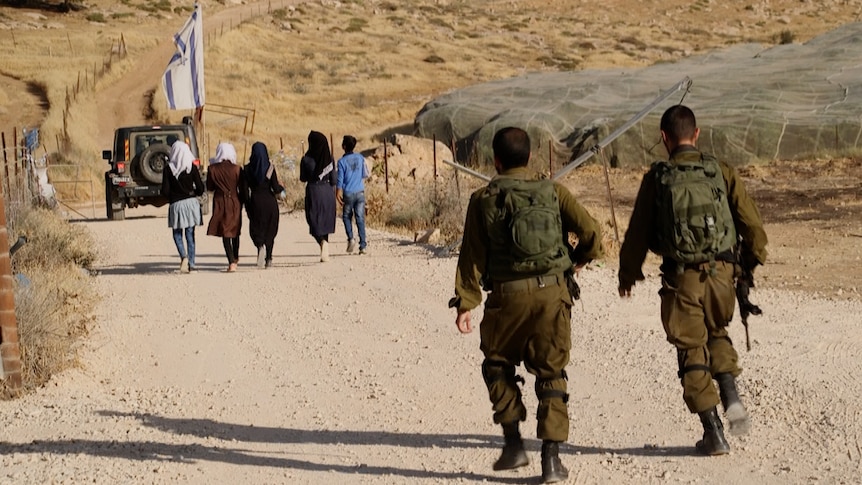 Palestinian teenagers walk to school accompanied by an armoured vehicle and two soldiers.