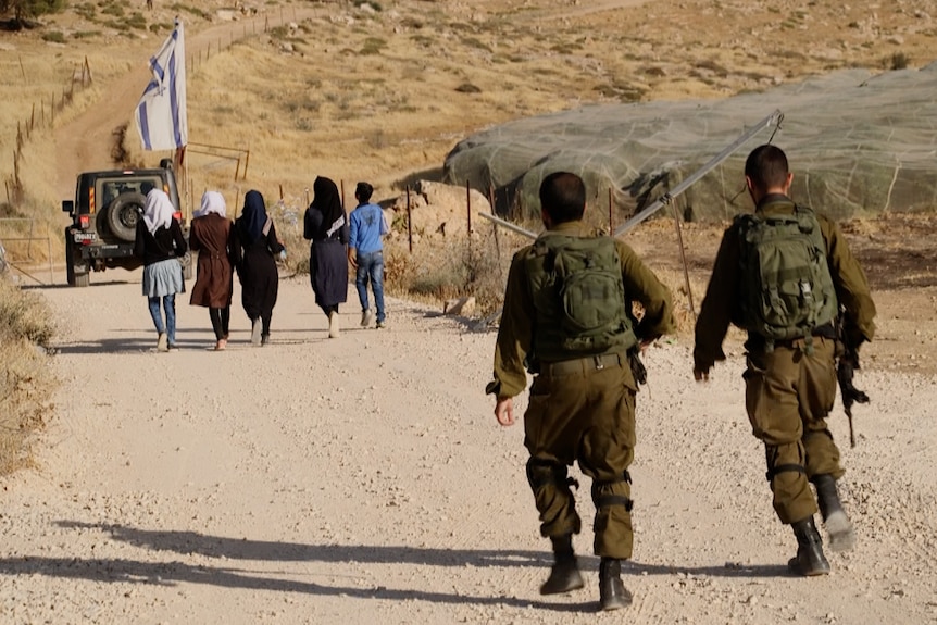 Palestinian teenagers walk to school accompanied by an armoured vehicle and two soldiers.