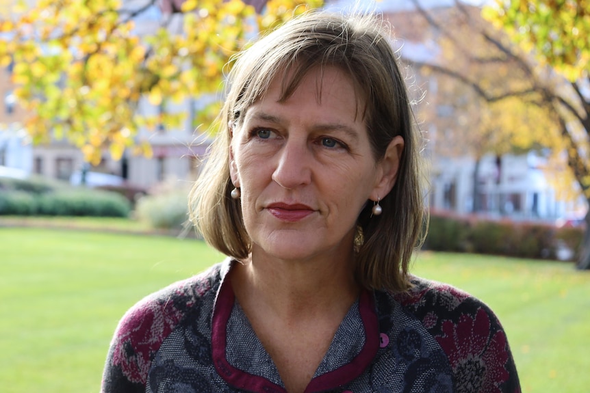 A woman stands on a green lawn with out of focus heritage buildings and autumn leaves in the background