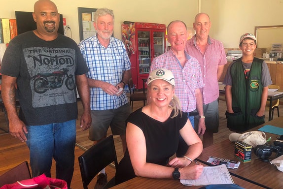 Senator Bridget McKenzie sits at a wooden table about to sign papers with a grop of men and  a young boy behind her