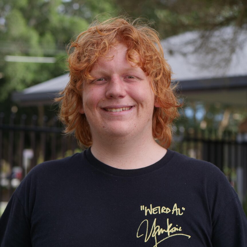 young boy with orange hair looks at the camera. He wears a white t shirt with the word 'mustang' on it. 