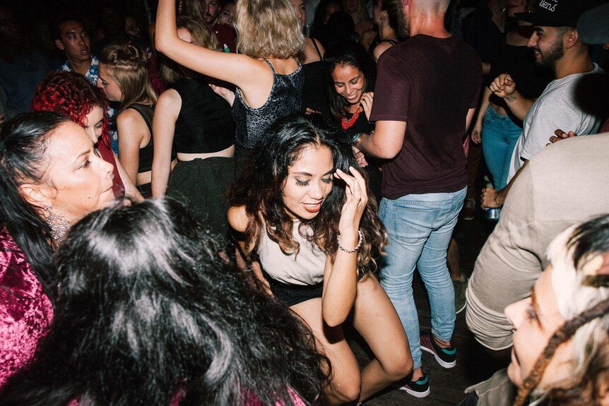 In a crowd of people at a party a woman is dancing, she is crouching low as her friends watch on.