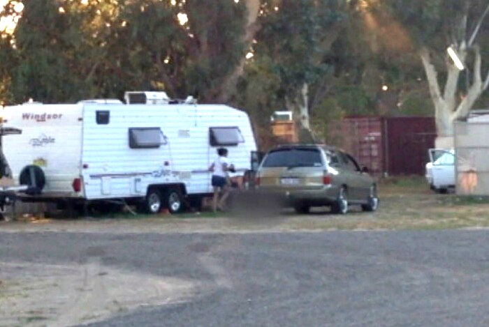 A station wagon parked next to a caravan with a woman in a white top next to a blurred out image of a man on the ground.