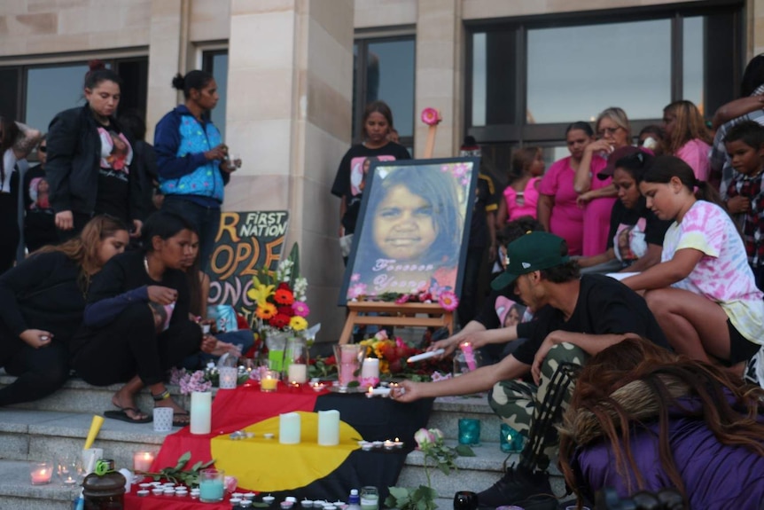 Dozens of people around a makeshift vigil on the steps of parliament house with flowers and lighting candles.