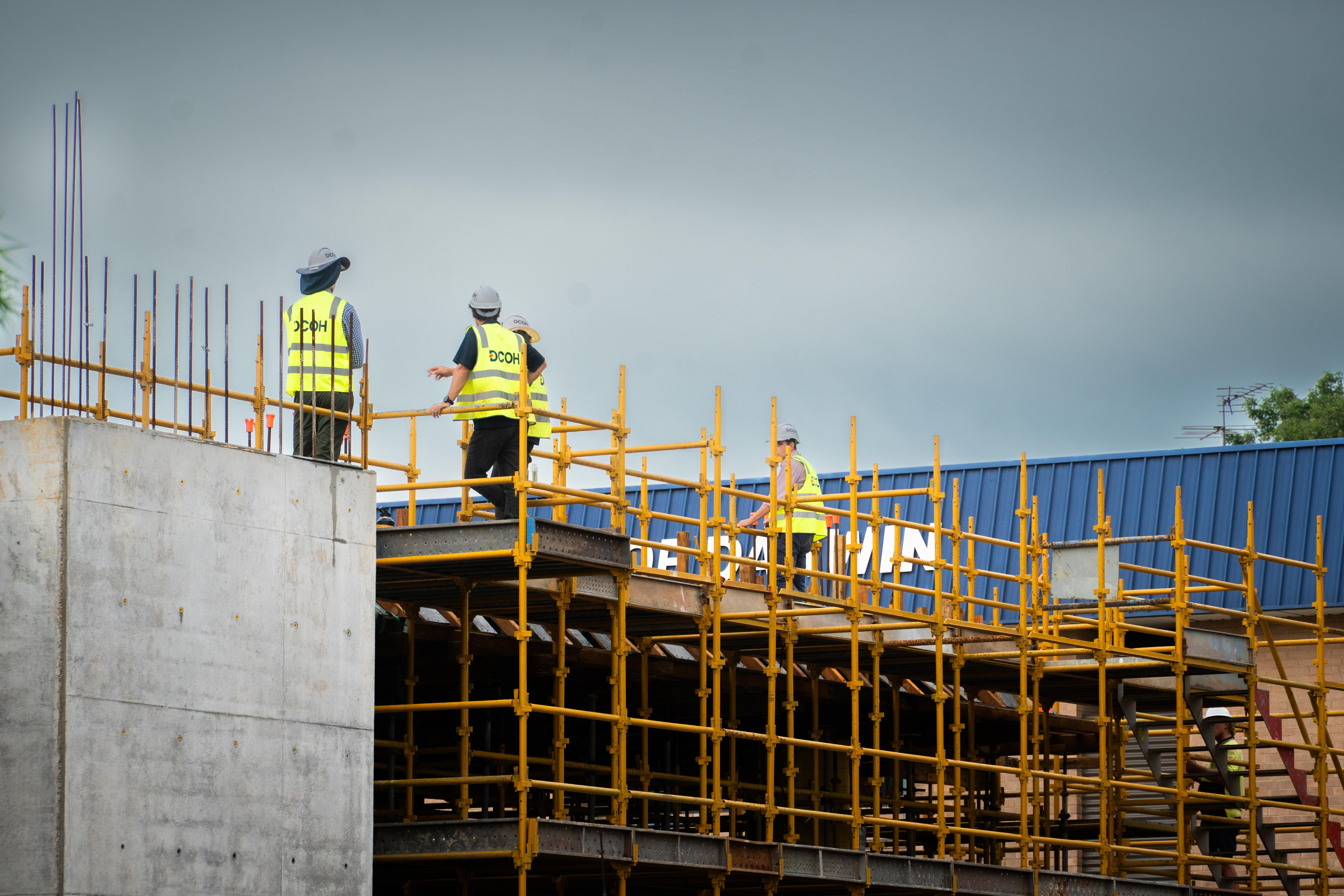 Pessoas usando capacetes e coletes de alta visibilidade em cima de um andaime em um canteiro de obras.