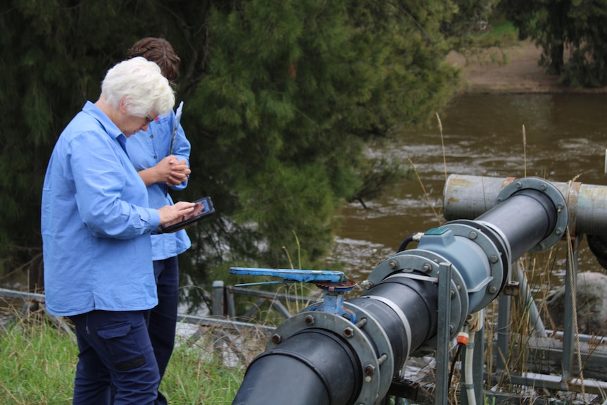 Two women with clipboards in blue government shirts inspect an irrigator water pump